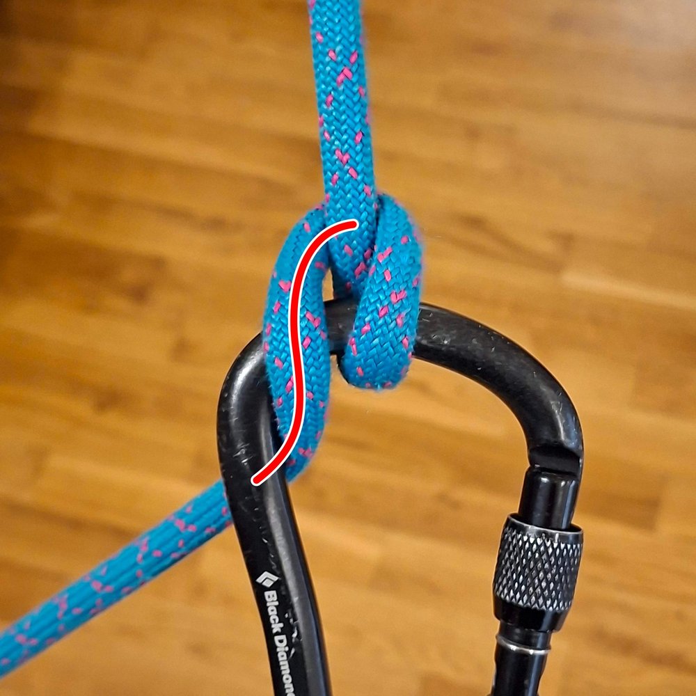 The Munter hitch on a carabiner with the brake strand leaving to the side. A red line traces a
                  pronounced S-shaped helical bend over the carabiner.