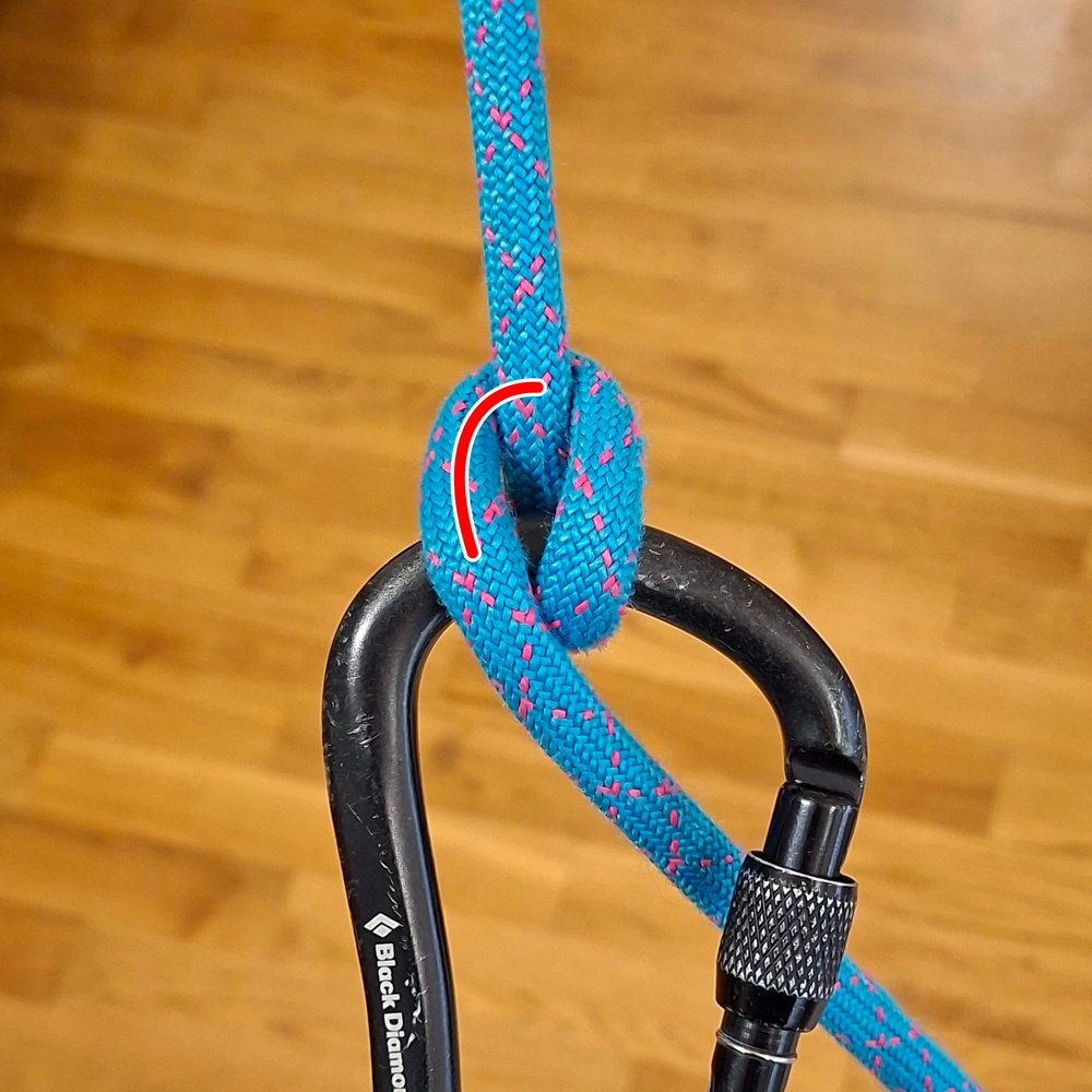 The Munter hitch with the brake strand leaving downwards across the carabiner. A short red line
                  marks a smaller helical bend at the top of the knot.
