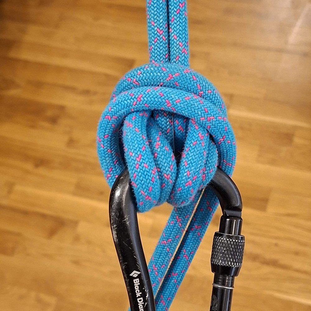 Two strands of a blue rope tied in a bulky Munter hitch on a carabiner, with the brake strands wrapped
              over the top of the knot.